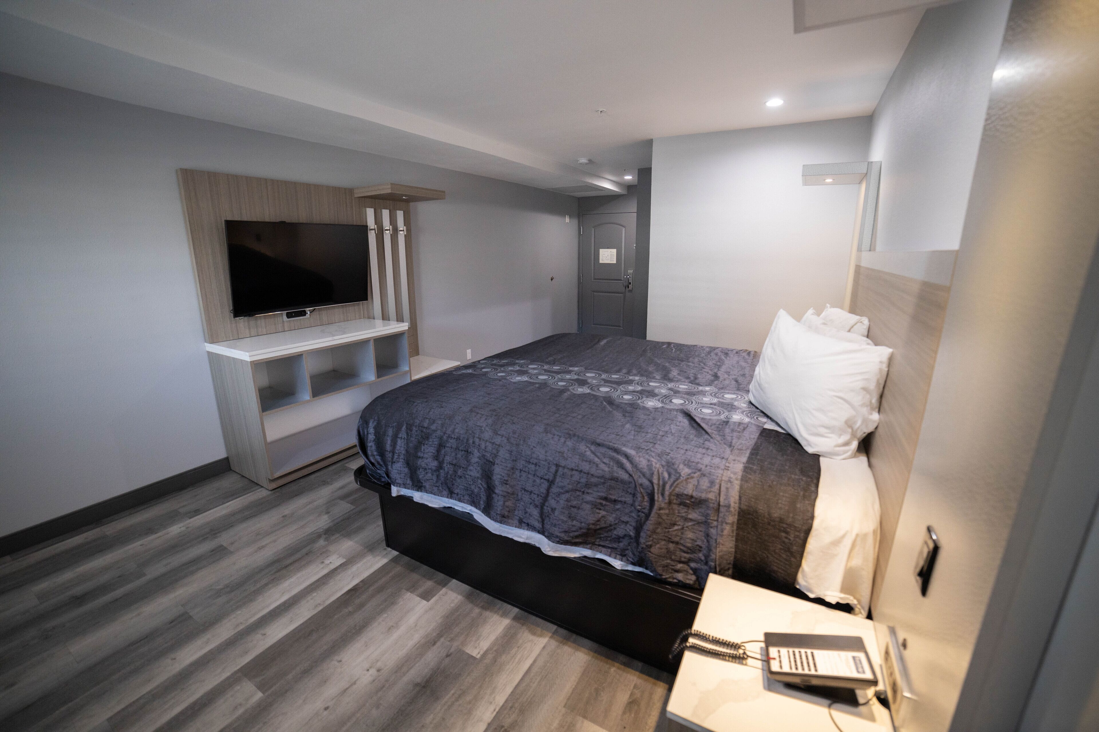 Une chambre moderne avec un grand lit, un téléviseur fixé sur une étagère en bois, une table de chevet avec une lampe et une porte dotée d’une poignée.