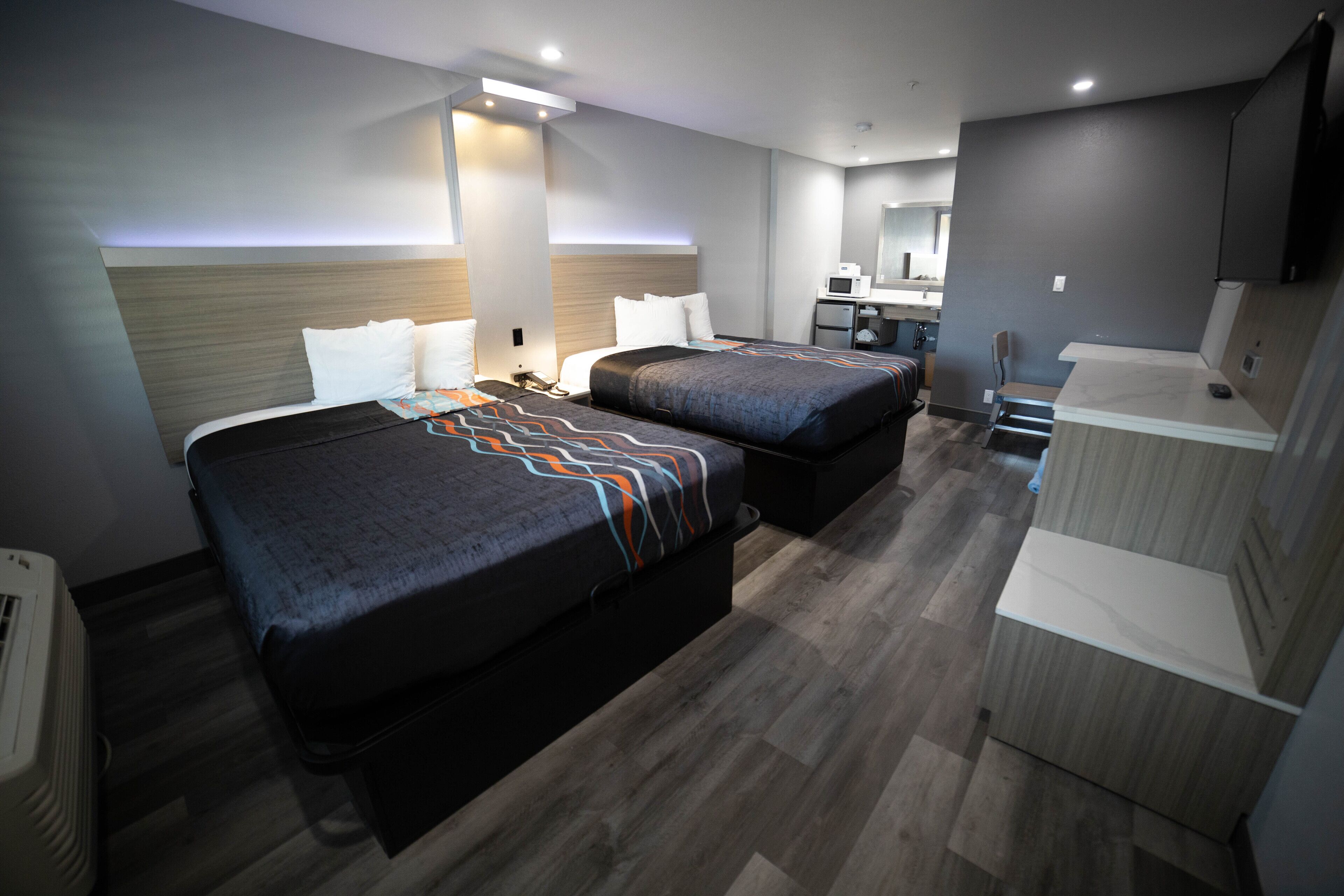 A hotel room with two beds, a desk, a chair, a microwave, and a small kitchenette.