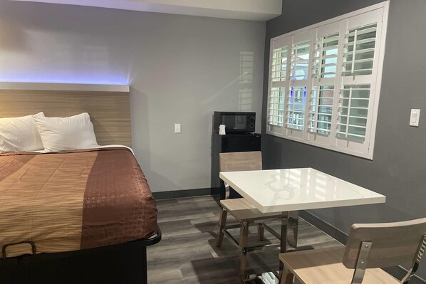 A hotel room with a bed, a table, a chair, a microwave, and a window with blinds.