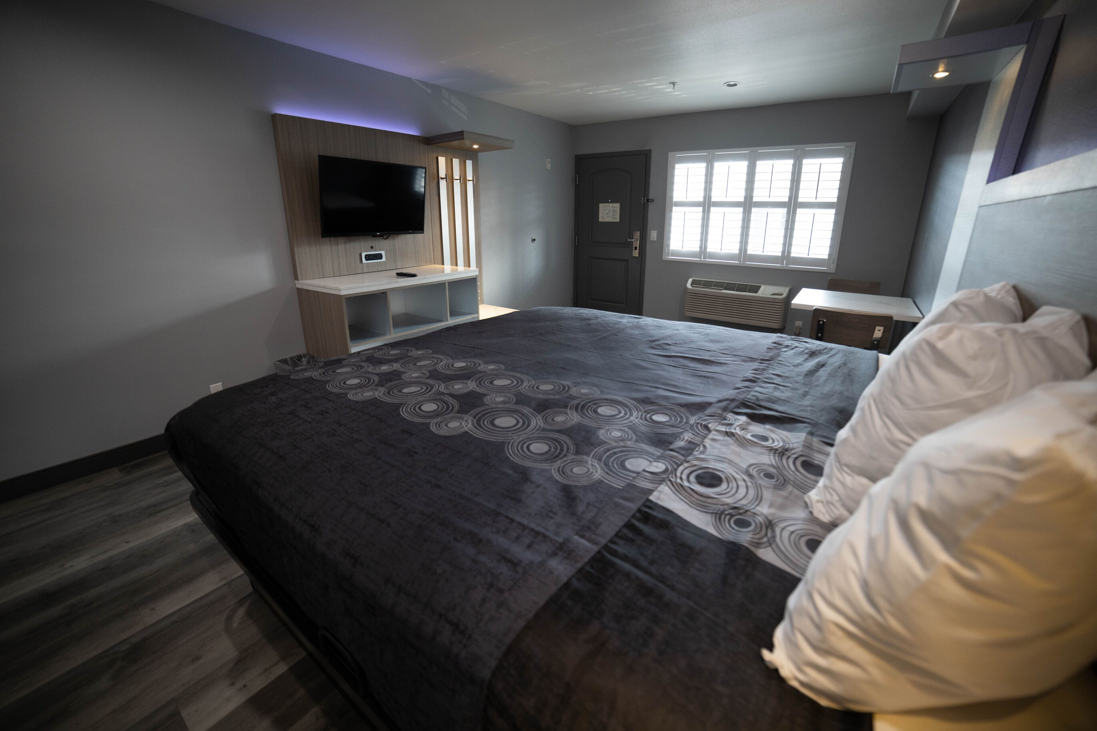 A modern bedroom with a large bed, a TV mounted on the wall, a desk, and a window.