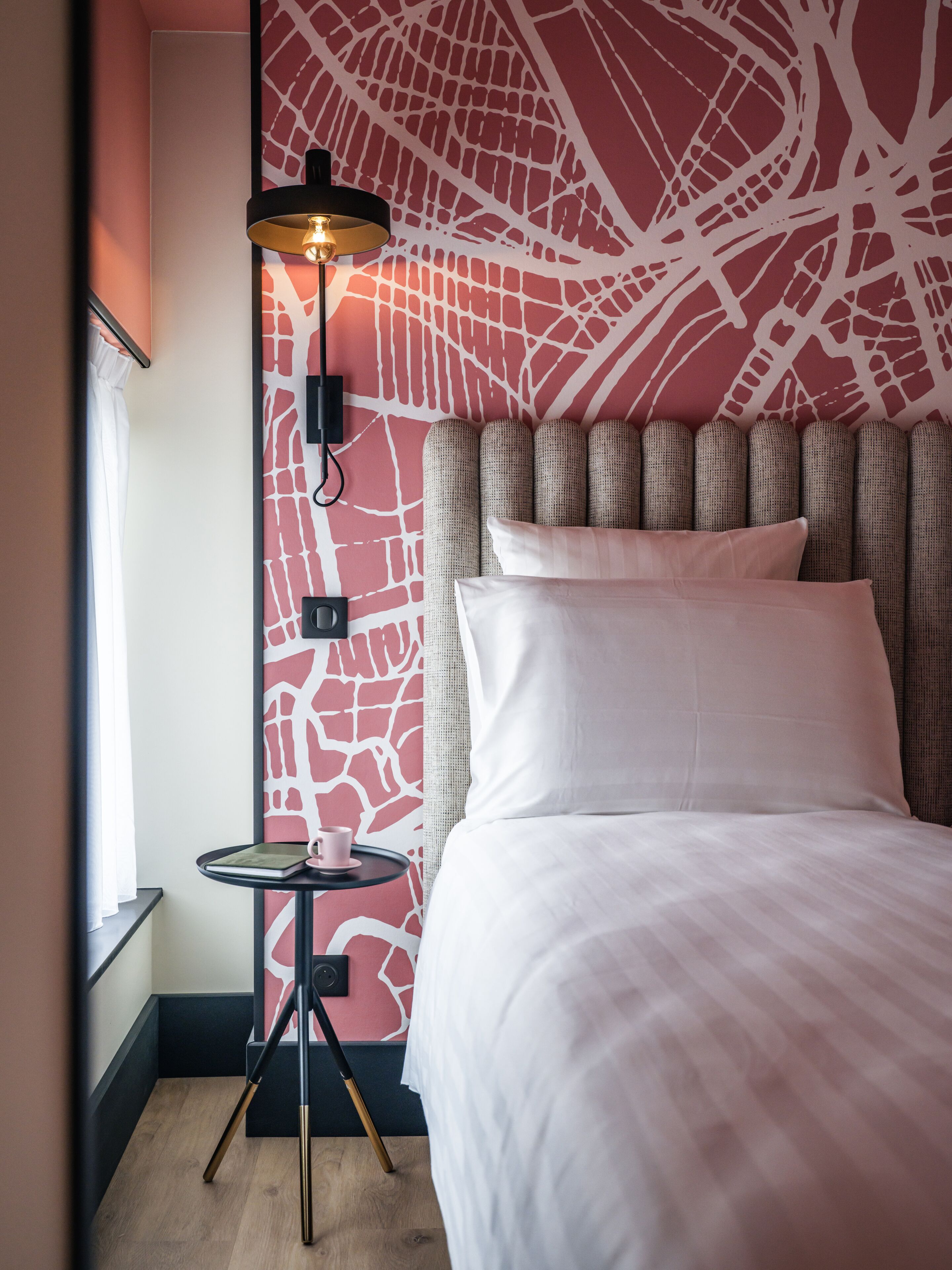 A hotel room with a bed, a bedside table with a lamp, and a wall with a red abstract design.