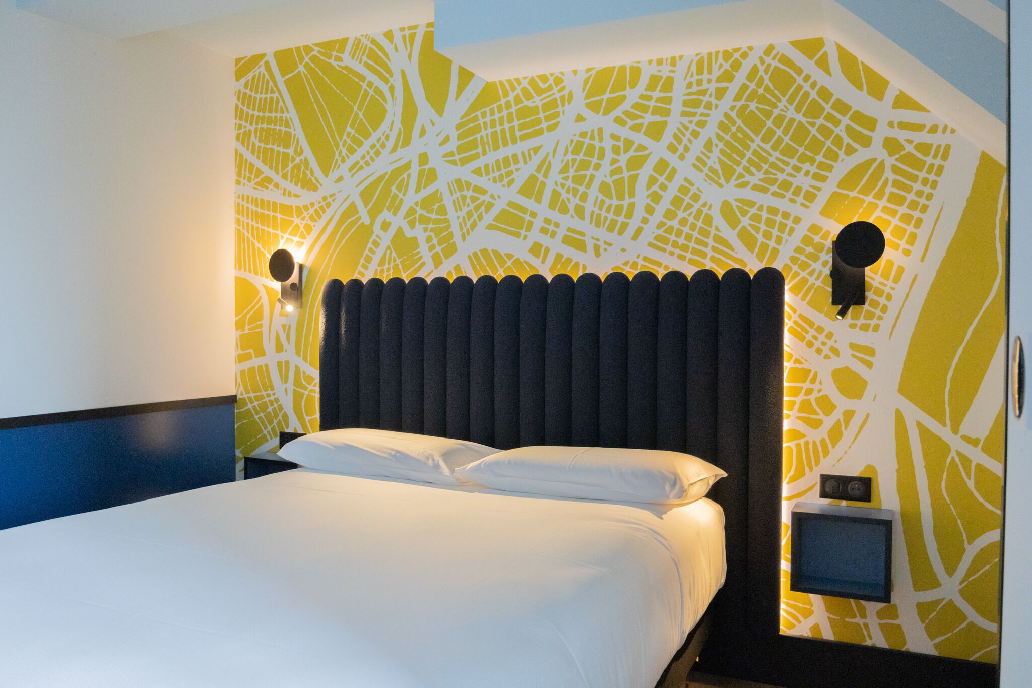 A modern hotel room with a large bed, a cityscape wall design, and two wall-mounted light fixtures.