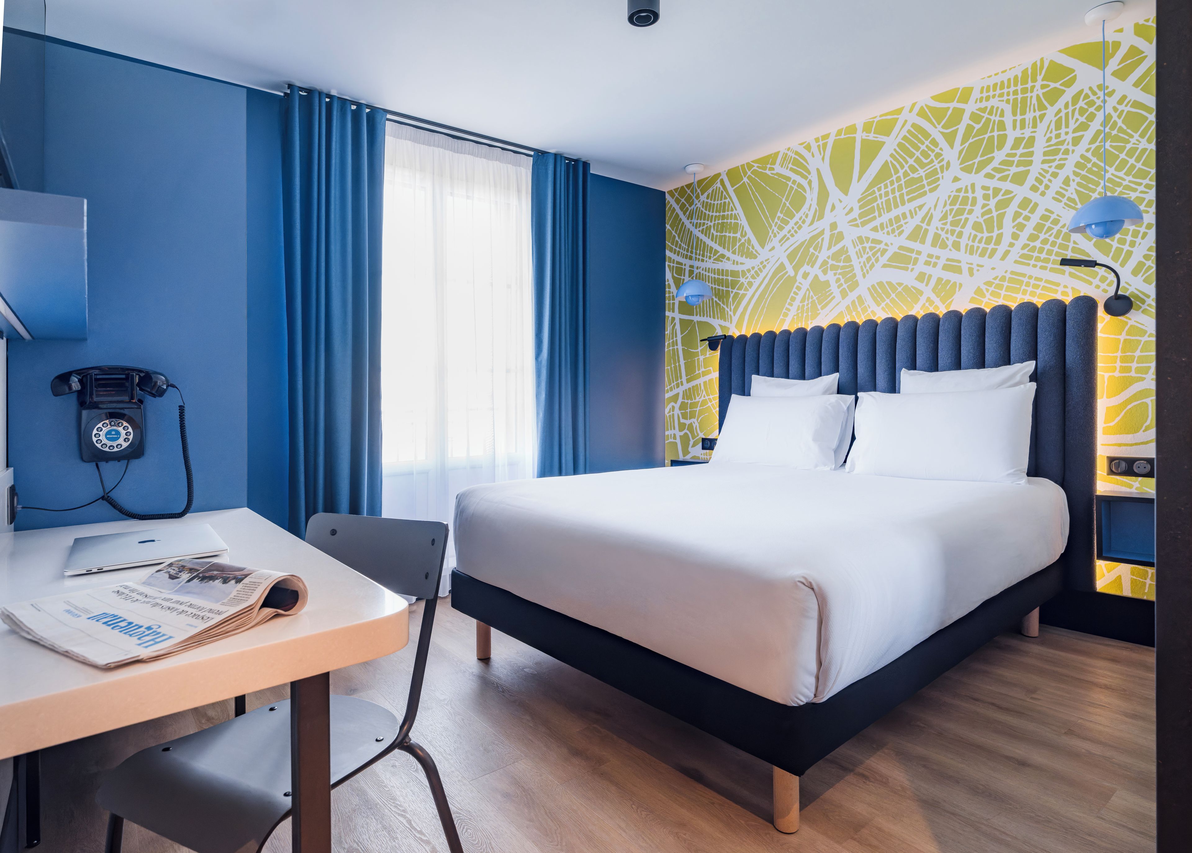 A hotel room with a bed, a desk with a telephone, and a window with curtains.