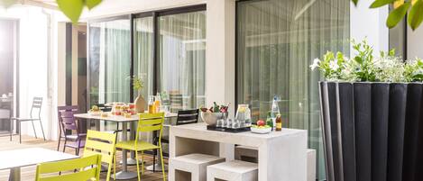 An outdoor dining area with a table set for two, surrounded by yellow chairs, and a planter with flowers to the right.
