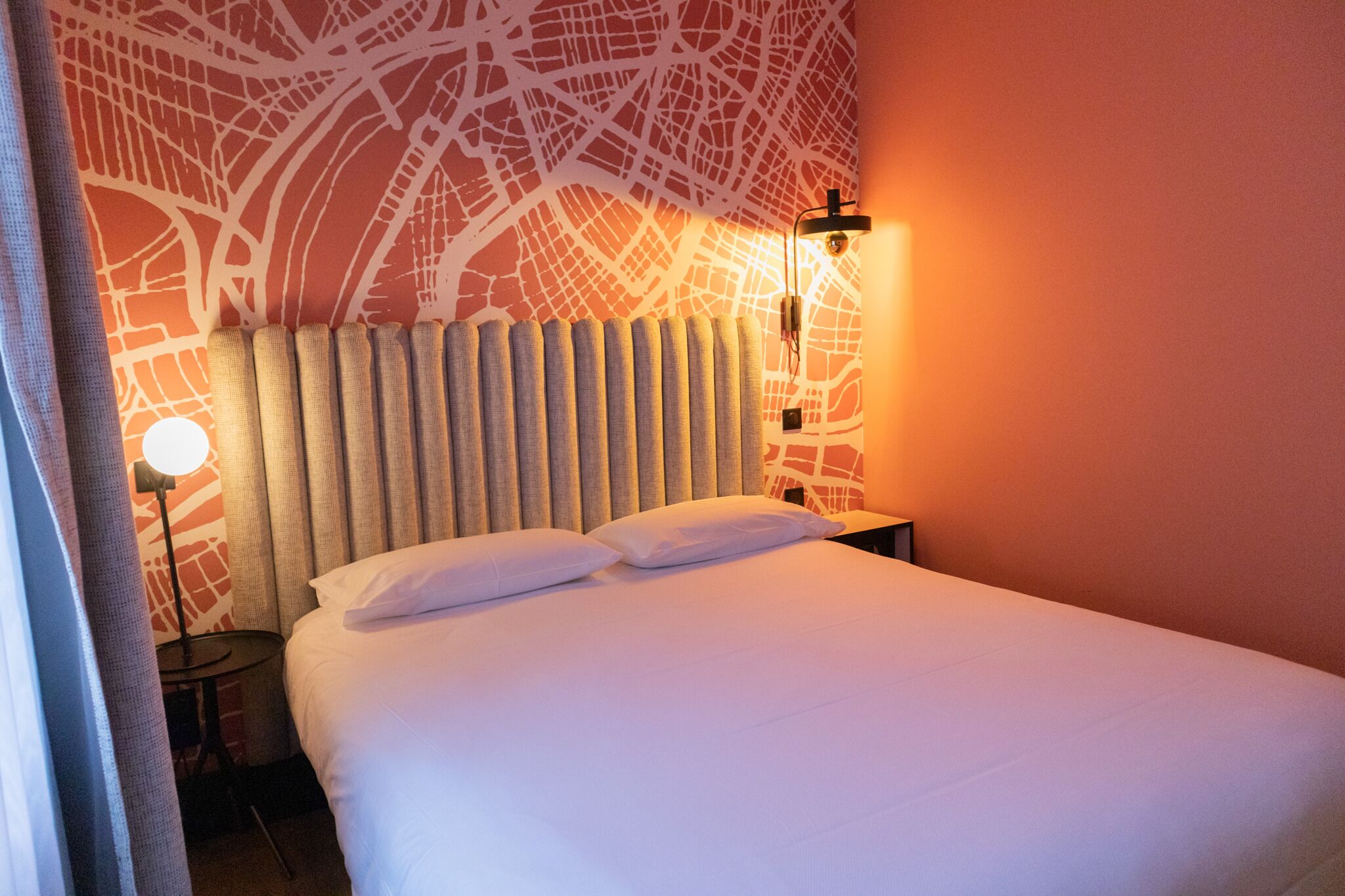 Un lit bien fait, avec du linge de lit blanc et des oreillers, une tête de lit  à lattes verticales, une lampe fixée au mur et un motif décoratif sur le mur.