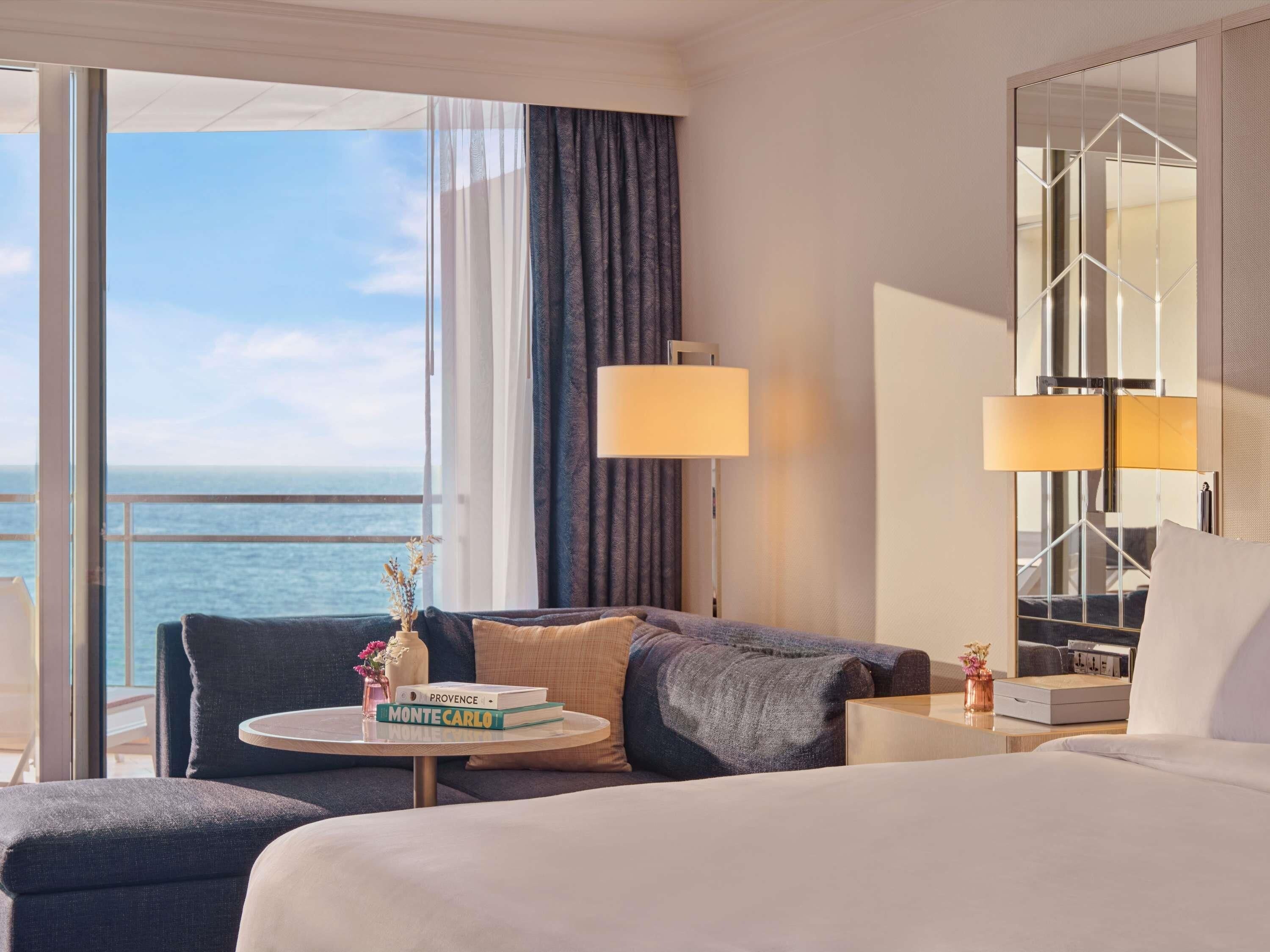 A hotel room with a bed, a sofa, a small table with books, a lamp, and a view of the sea.