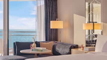 A hotel room with a bed, a sofa, a small table with books, a lamp, and a view of the sea.
