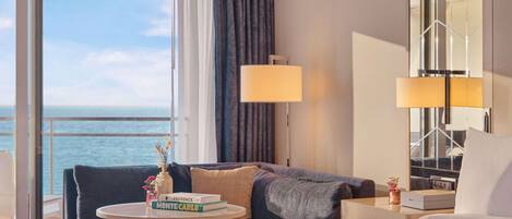 A hotel room with a bed, a sofa, a small table with books, a lamp, and a view of the sea.