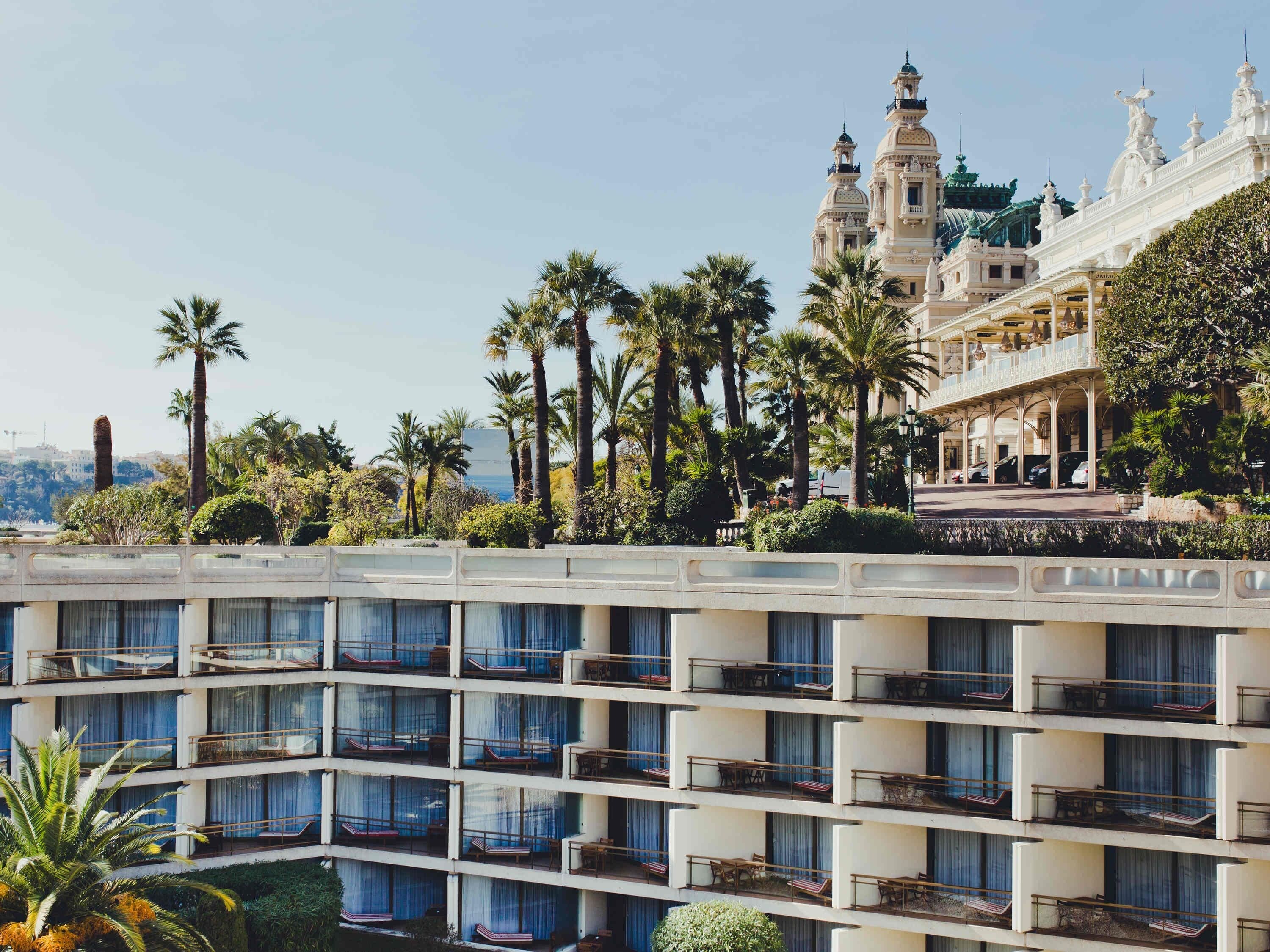Ein Hotel mit mehreren Balkonen, Palmen und einem im Hintergrund sichtbaren Gebäude mit aufwendiger Architektur.