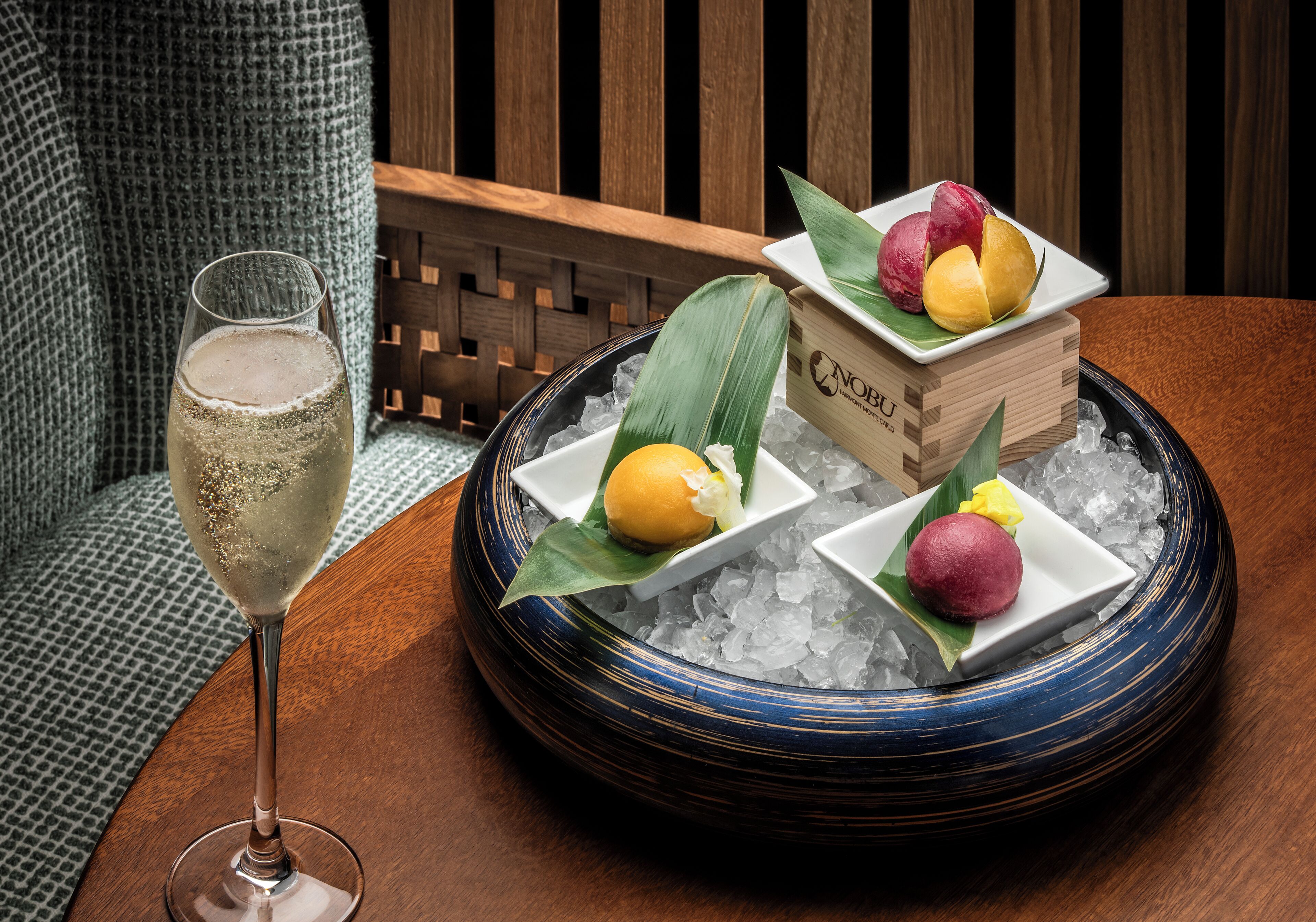 A table with a glass of a light-colored beverage, a blue-rimmed bowl with ice, and a wooden box containing three different types of fruit on a wooden surface.