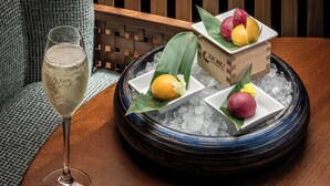 Una mesa con un vaso de una bebida de color claro, un cuenco con borde azul y hielo, y una caja de madera que contiene tres tipos diferentes de fruta.
