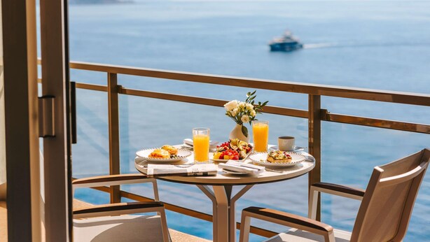 Un tavolo da balcone con la colazione, un vaso di fiori e vista sul mare.