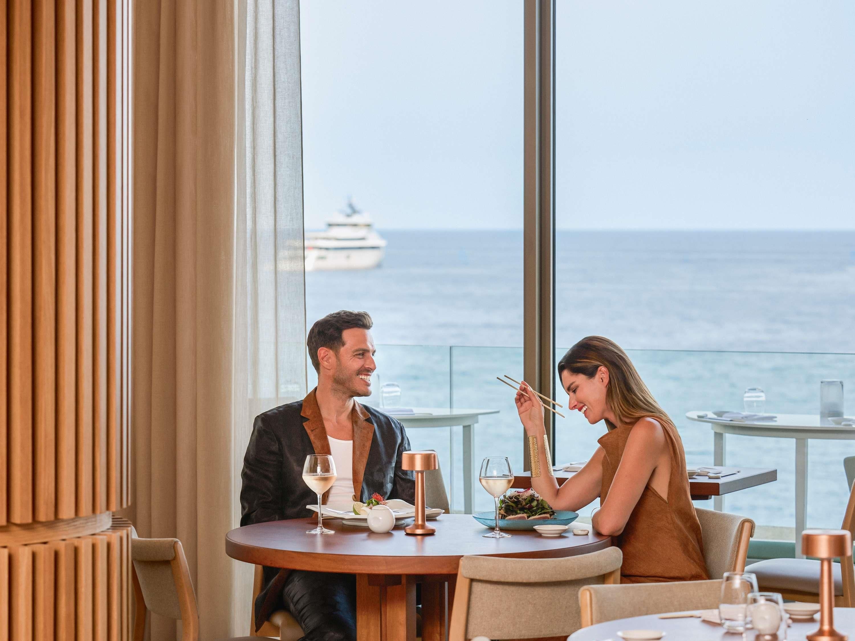 A dining area with a view of the sea and a yacht, featuring a round table with a glass of wine and a plate of food.