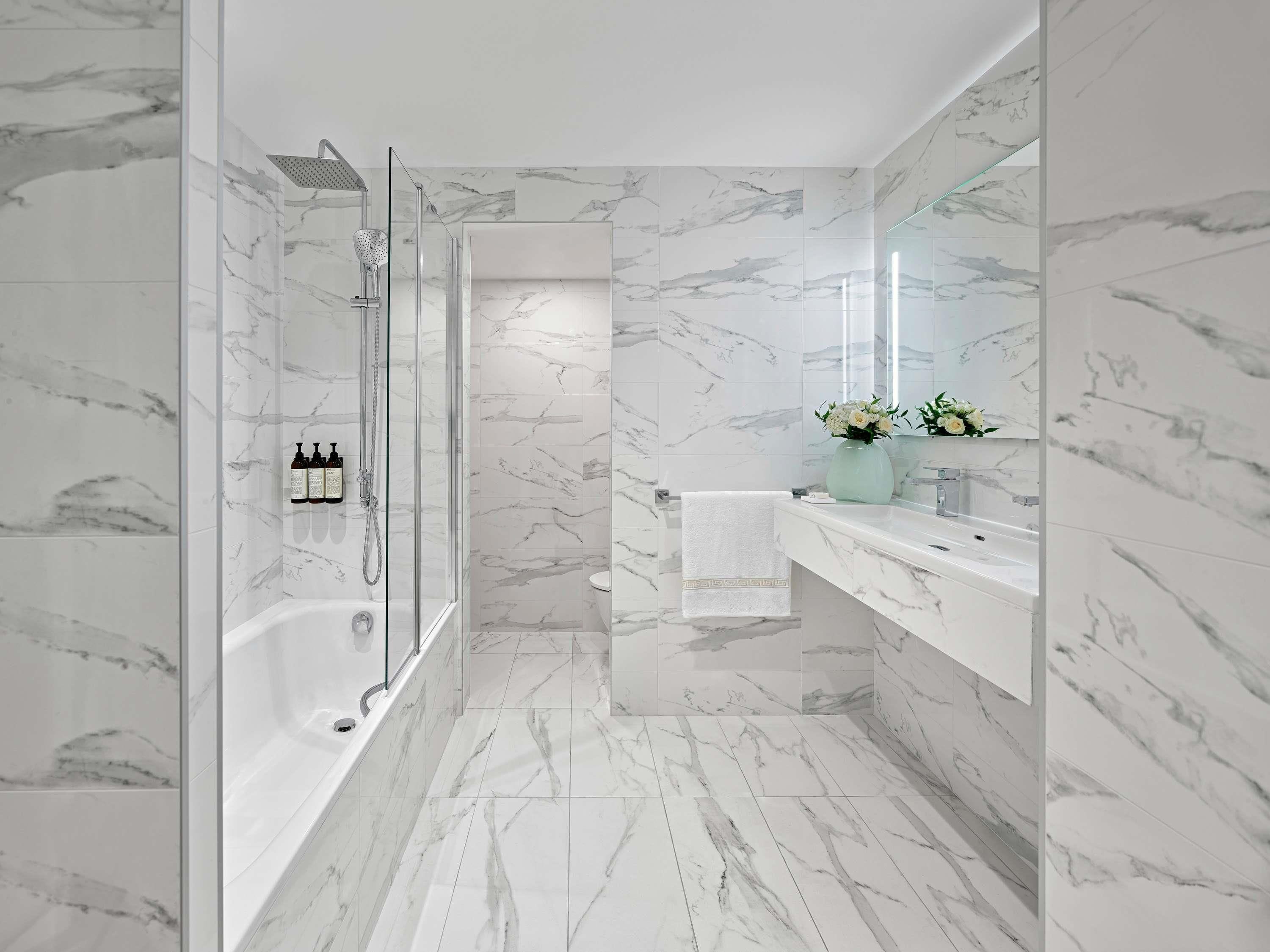 A modern bathroom with a glass-enclosed shower, a marble-tiled floor, and a vanity with a mirror and a vase of flowers.