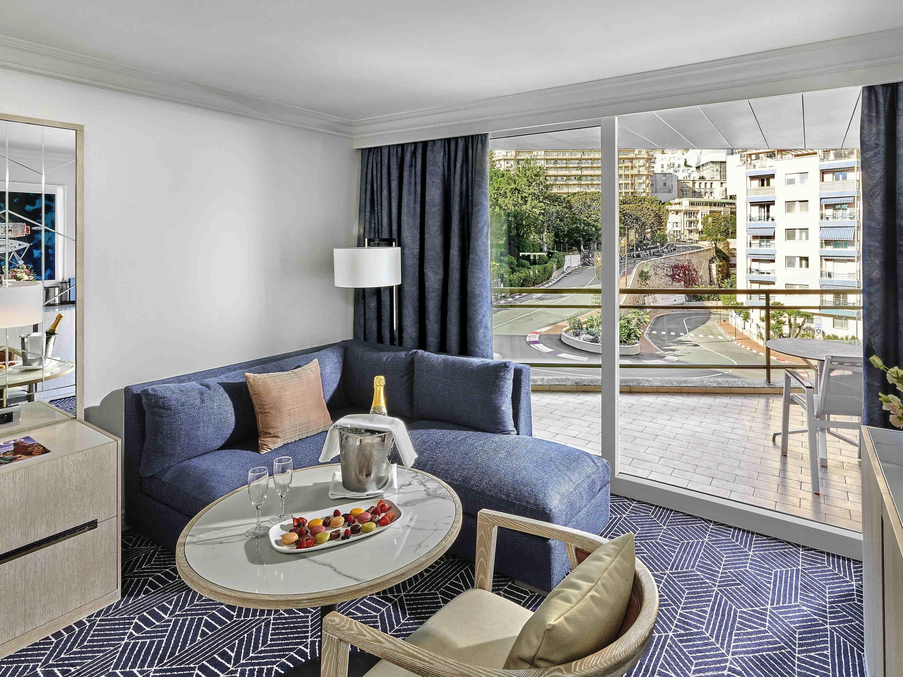 A modern hotel room with a sofa, a small round table with a bottle of champagne, a tray of snacks, and a view of the cityscape through large windows.