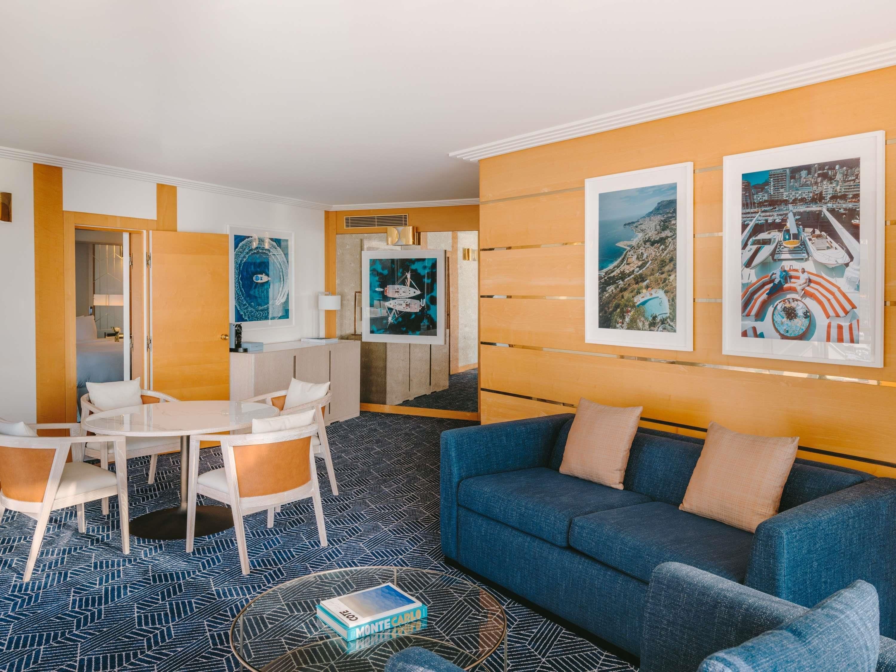 A modern hotel room with a blue sofa, a dining area with a white table and chairs, and artwork on the walls.