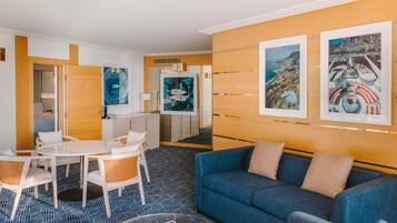A modern hotel room with a blue sofa, a dining area with a white table and chairs, and artwork on the walls.