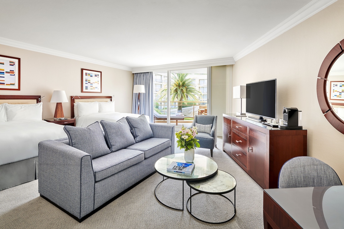 A hotel room with a bed, grey sofa, round coffee table, TV, and a view of the outdoors.