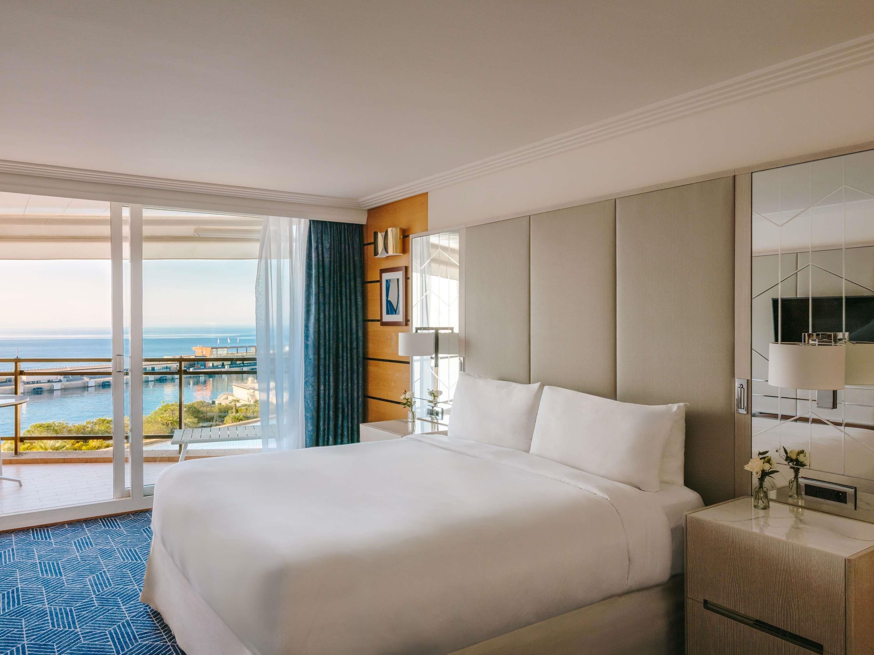 A hotel room with a large bed, a bedside table, a mirror, and a view of the sea.