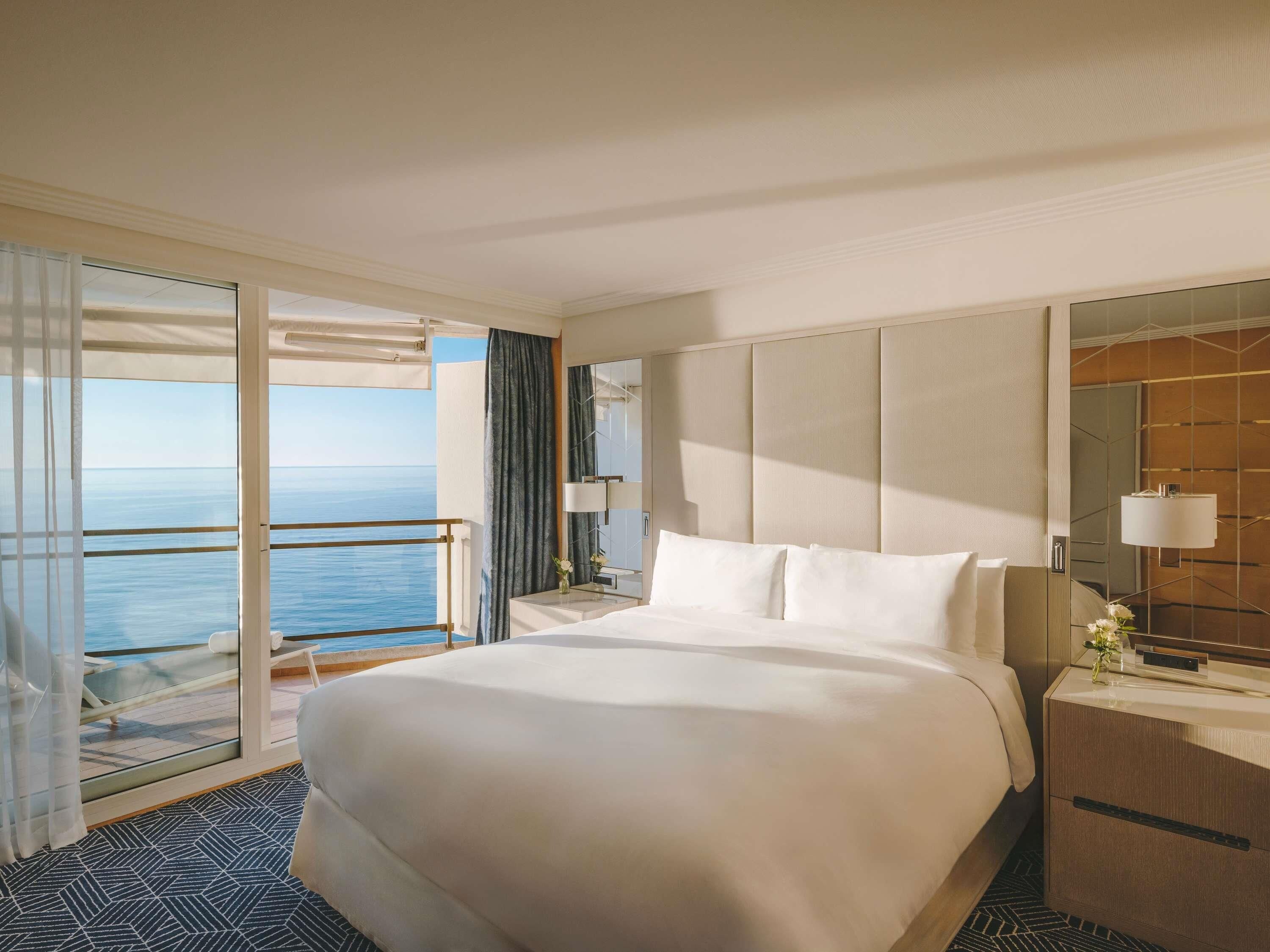 A hotel room with a large bed, a bedside table, a mirror, and a view of the sea.