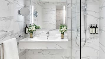 Et moderne badeværelse med marmorvægge, en glasafskærmet brusekabine, en hvid vask med to spejle og toiletartikler på væggen.