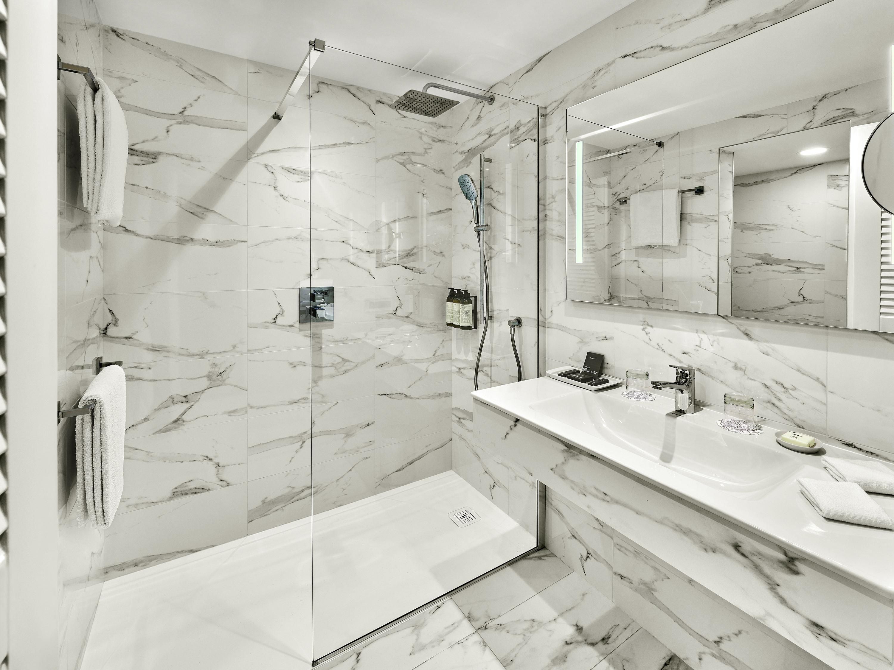 A modern bathroom with a glass-enclosed shower, a marble-tiled wall, a large mirror, and a white countertop with a sink and toiletries.