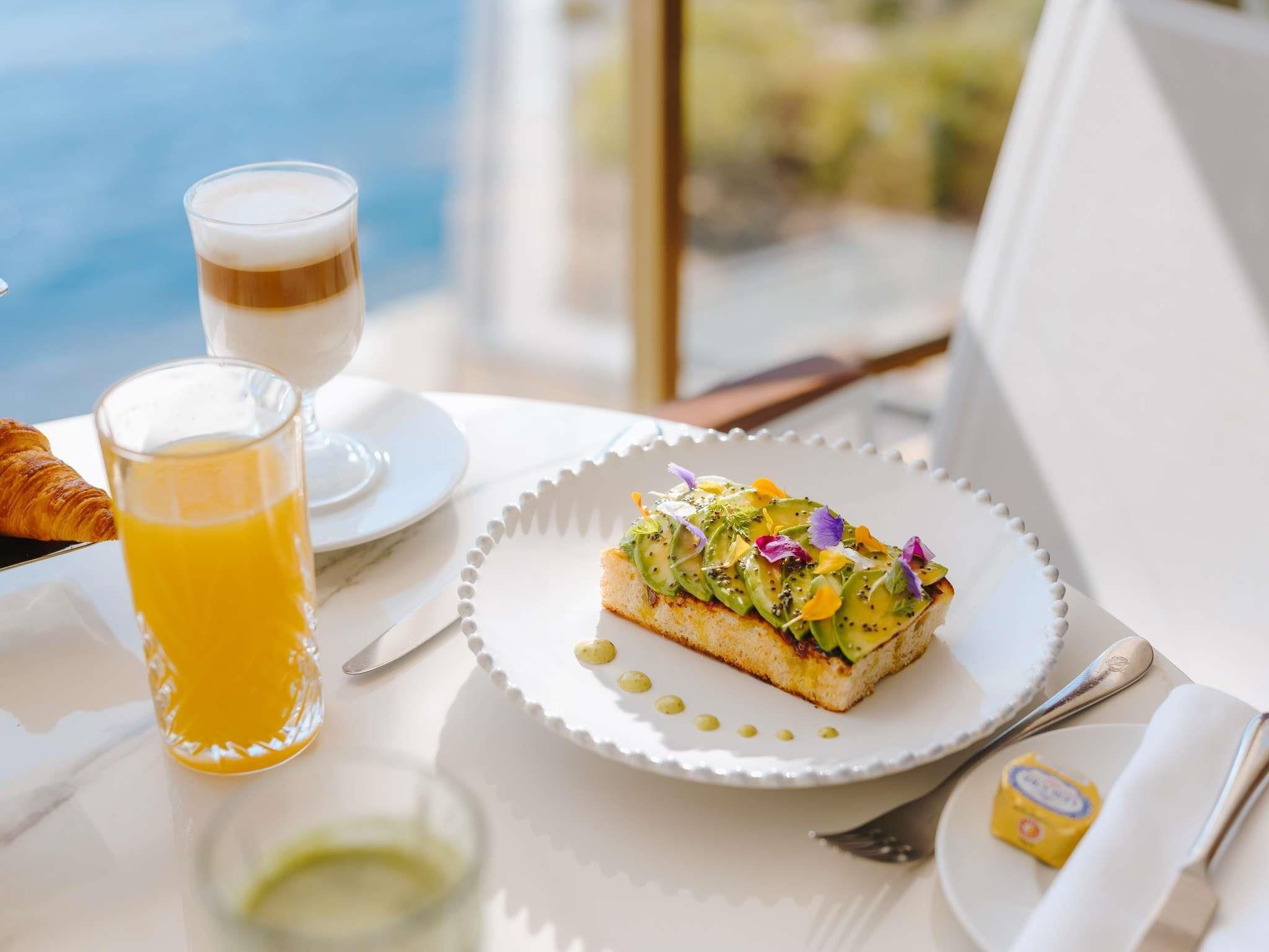 Desayuno con un croissant, un vaso de zumo de naranja y una tostada cubierta de fruta y vegetales, todo sobre un plato blanco.