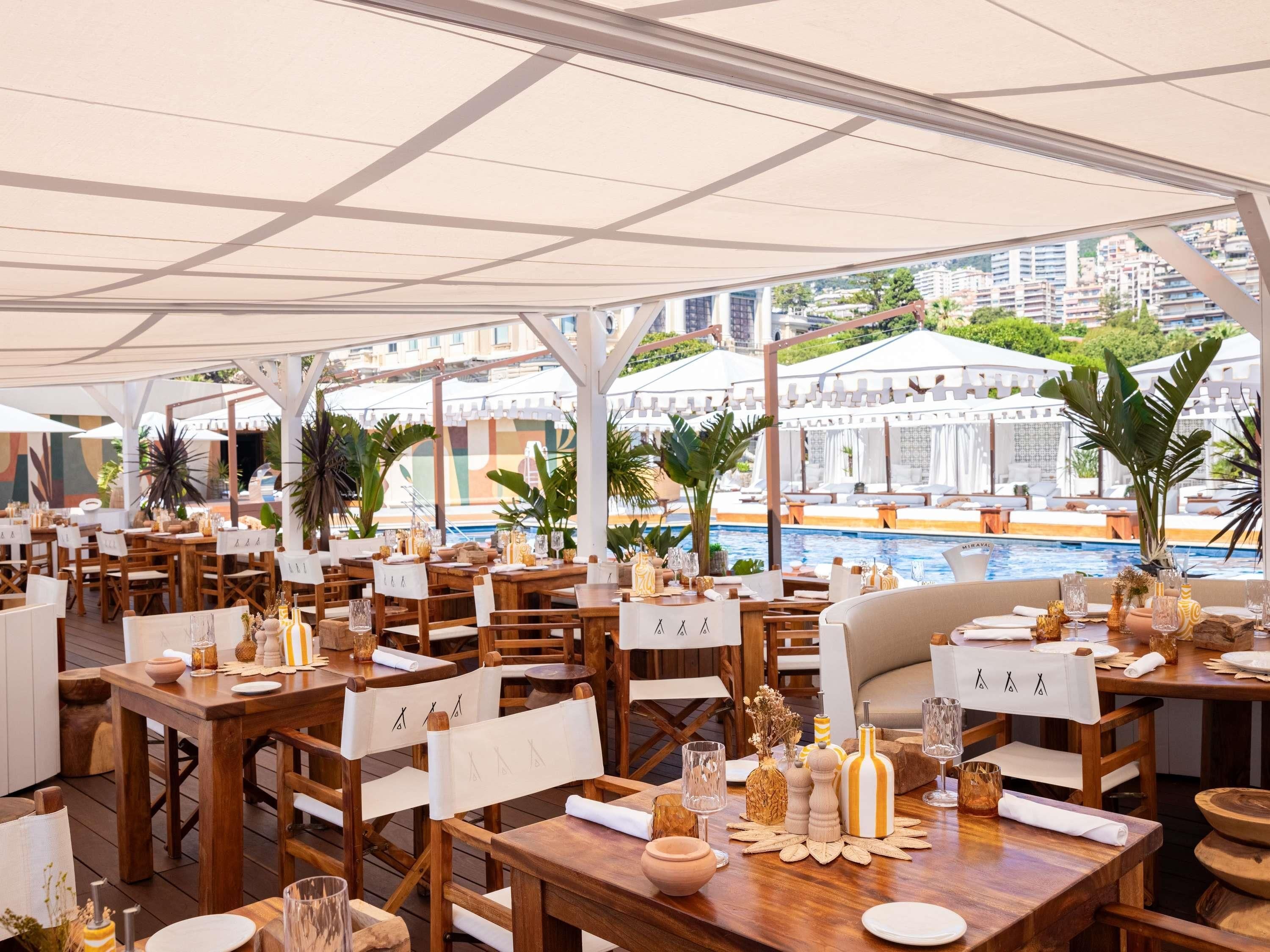 An outdoor dining area with wooden tables and chairs, surrounded by umbrellas, with a pool and buildings in the background.