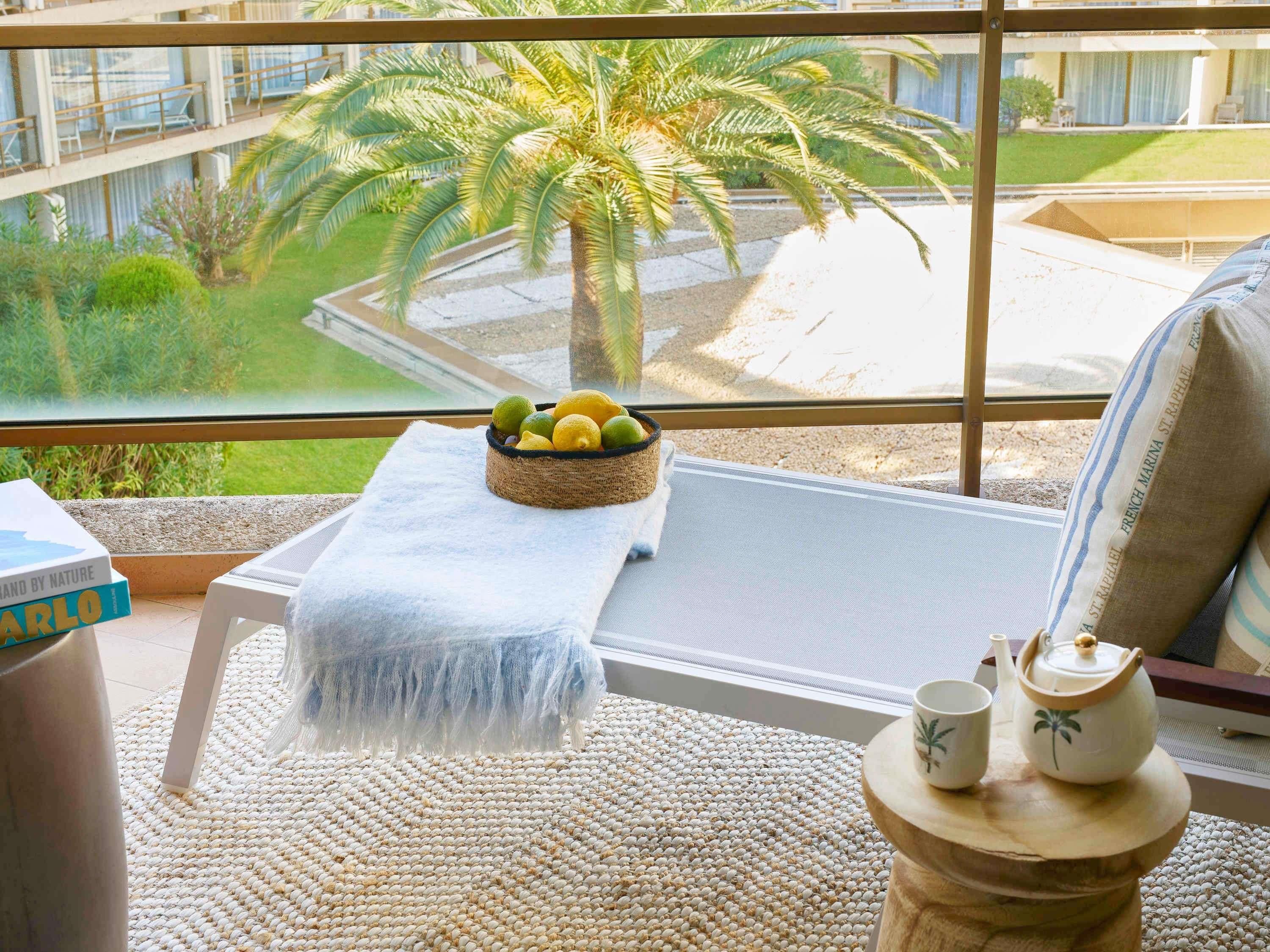 Un balcon agrémenté d’un tapis en fibres tressées, une petite table avec un saladier de fruits, une serviette blanche et une vue sur un palmier et un immeuble.