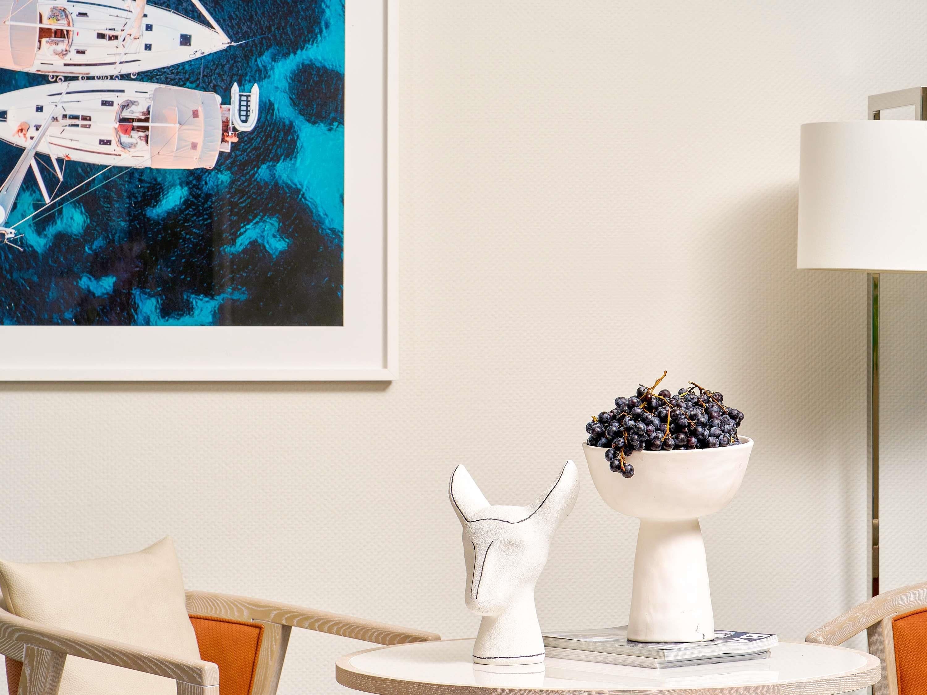 Ein Zimmer mit einem gerahmten Kunstwerk von Booten, einer weißen Keramikschale mit Trauben und einer weißen Keramikpferd-Figur auf einem kleinen Tisch.