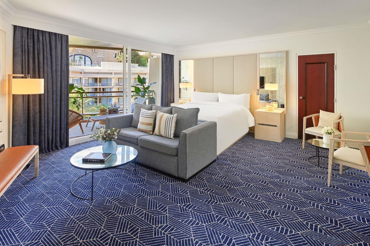 A hotel room with a bed, sofa, armchair, and a view of the outside.