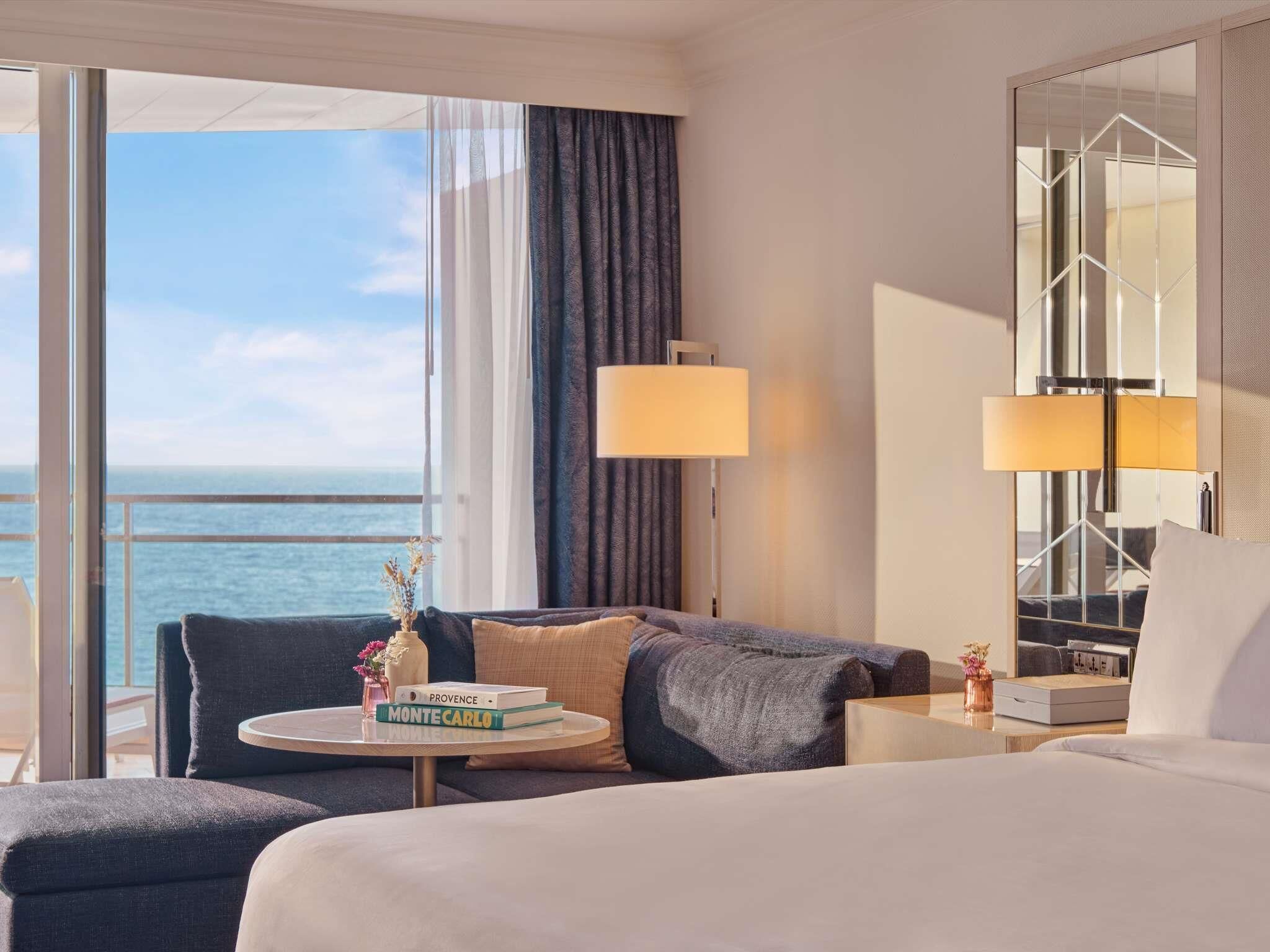 Une chambre d’hôtel avec un lit, une table ronde sur laquelle sont posés des livres, une lampe et une vue sur l’océan.