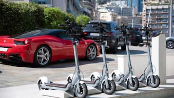 A row of electric scooters parked on a sidewalk with a red sports car and a black car in the background.