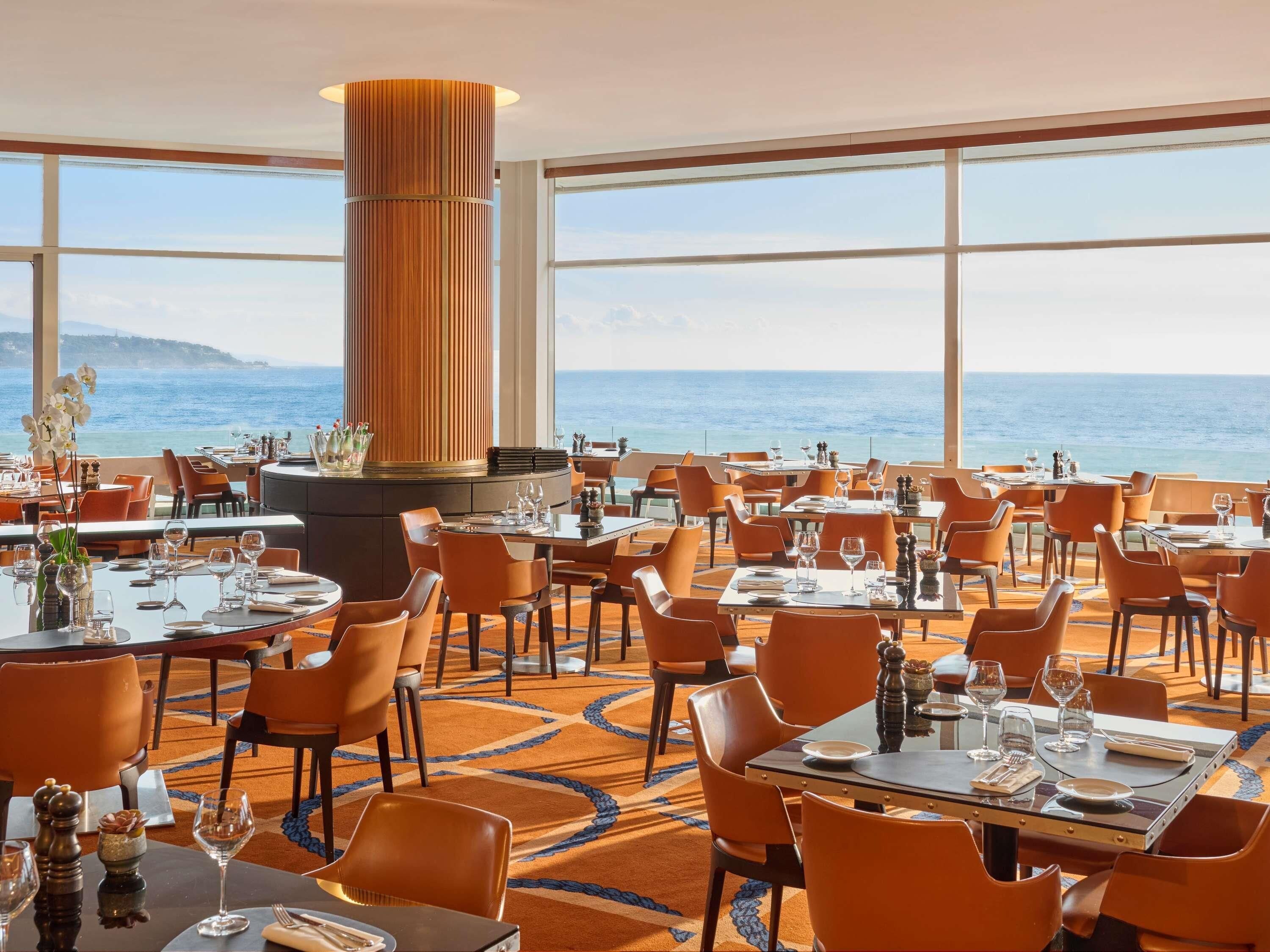 A spacious dining area with tables and chairs, large windows offering a view of the sea, and a central wooden column.