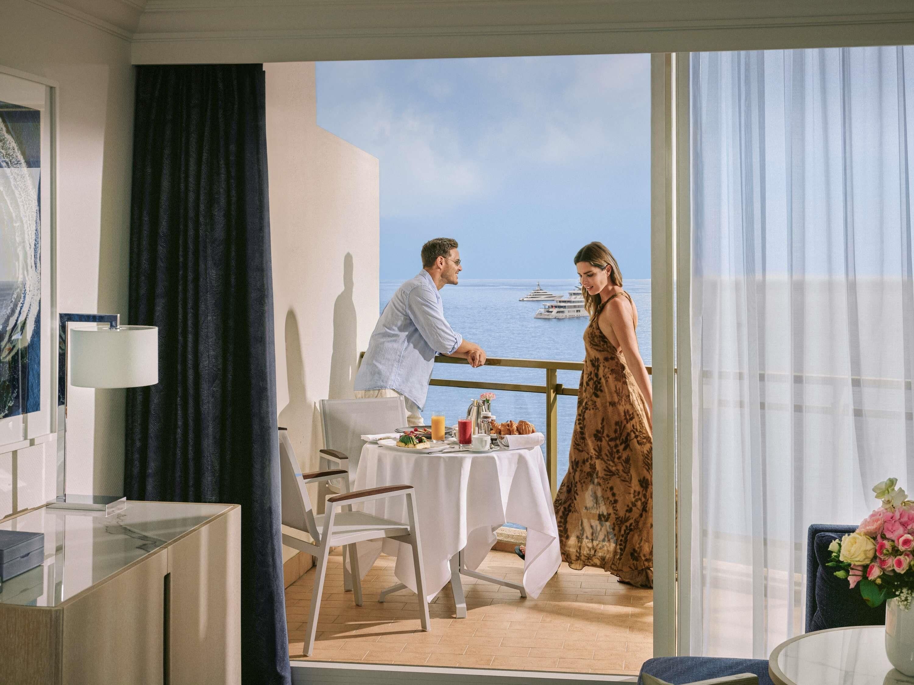 Una habitación con balcón que da al mar, una mesa de comedor con comida y vista a yates.