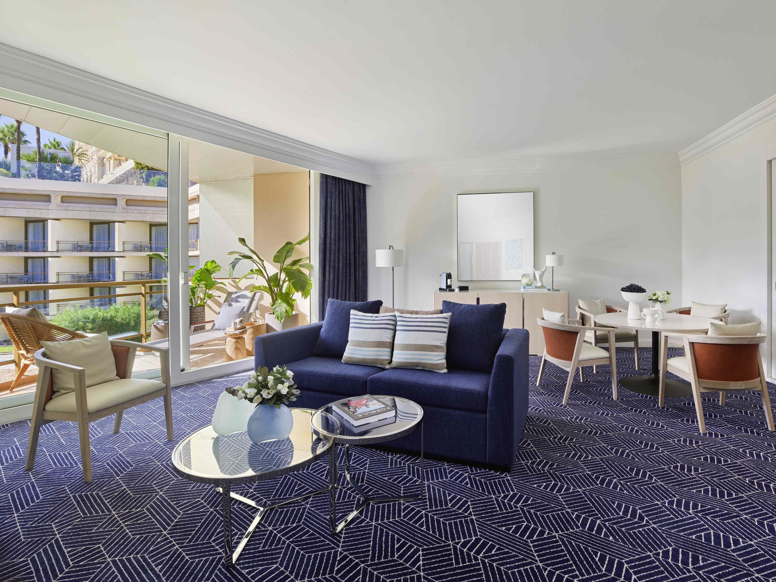 Une chambre d’hôtel équipée d’un canapé, d’une table basse, d’une table à manger, de chaises et d’une grande fenêtre donnant sur un immeuble et sur de la verdure.