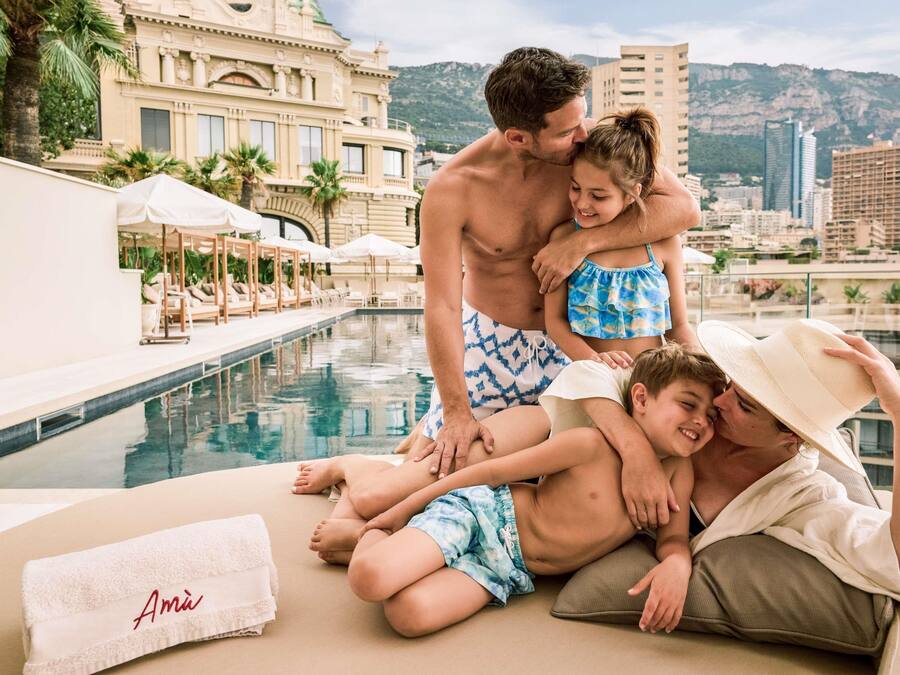 Escena junto a la piscina con una familia, una piscina, sillas de descanso y un edificio de arquitectura clásica al fondo.