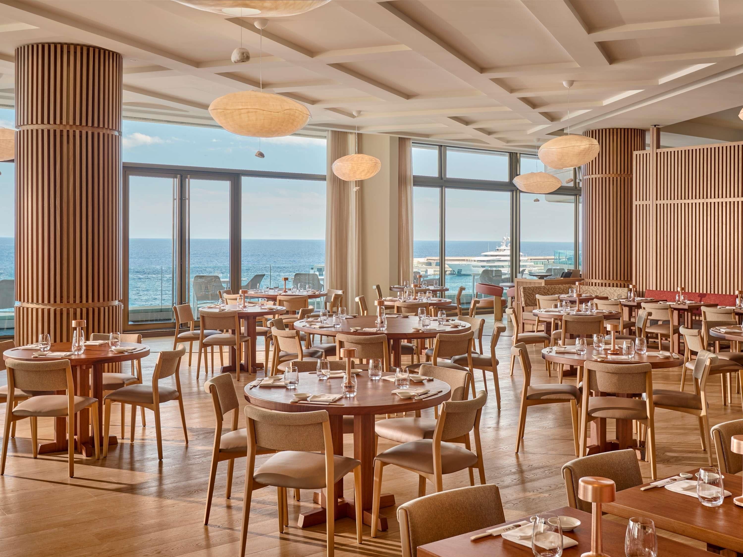 A spacious dining area with multiple tables and chairs, large windows offering a view of the sea and boats, and wooden paneling on the walls.