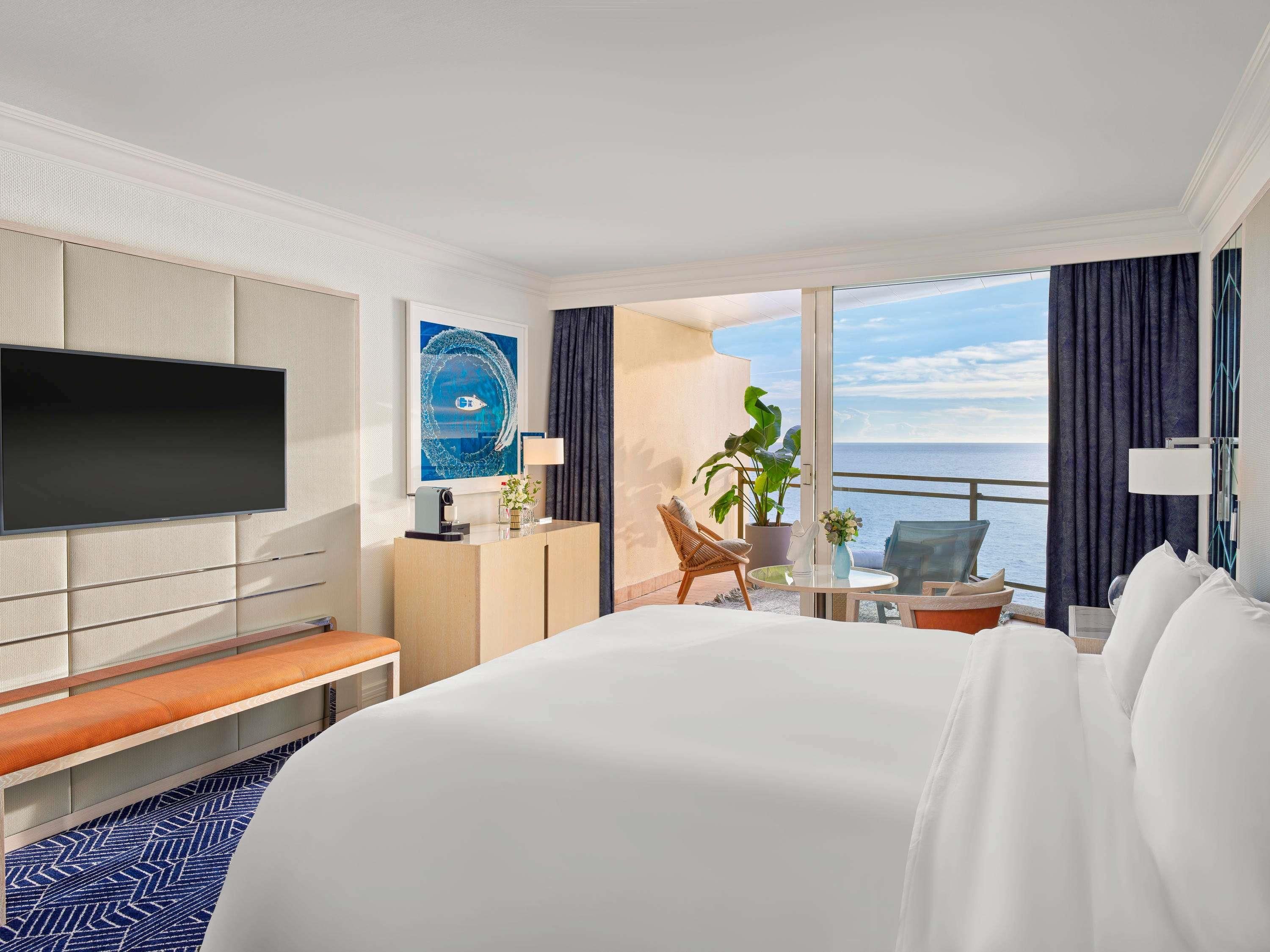 A hotel room with a large bed, a flat-screen TV mounted on the wall, a desk with a lamp, a chair, a potted plant, and a view of the ocean.