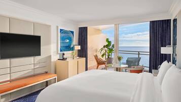 A hotel room with a large bed, a flat-screen TV mounted on the wall, a desk with a lamp, a chair, a potted plant, and a view of the ocean.