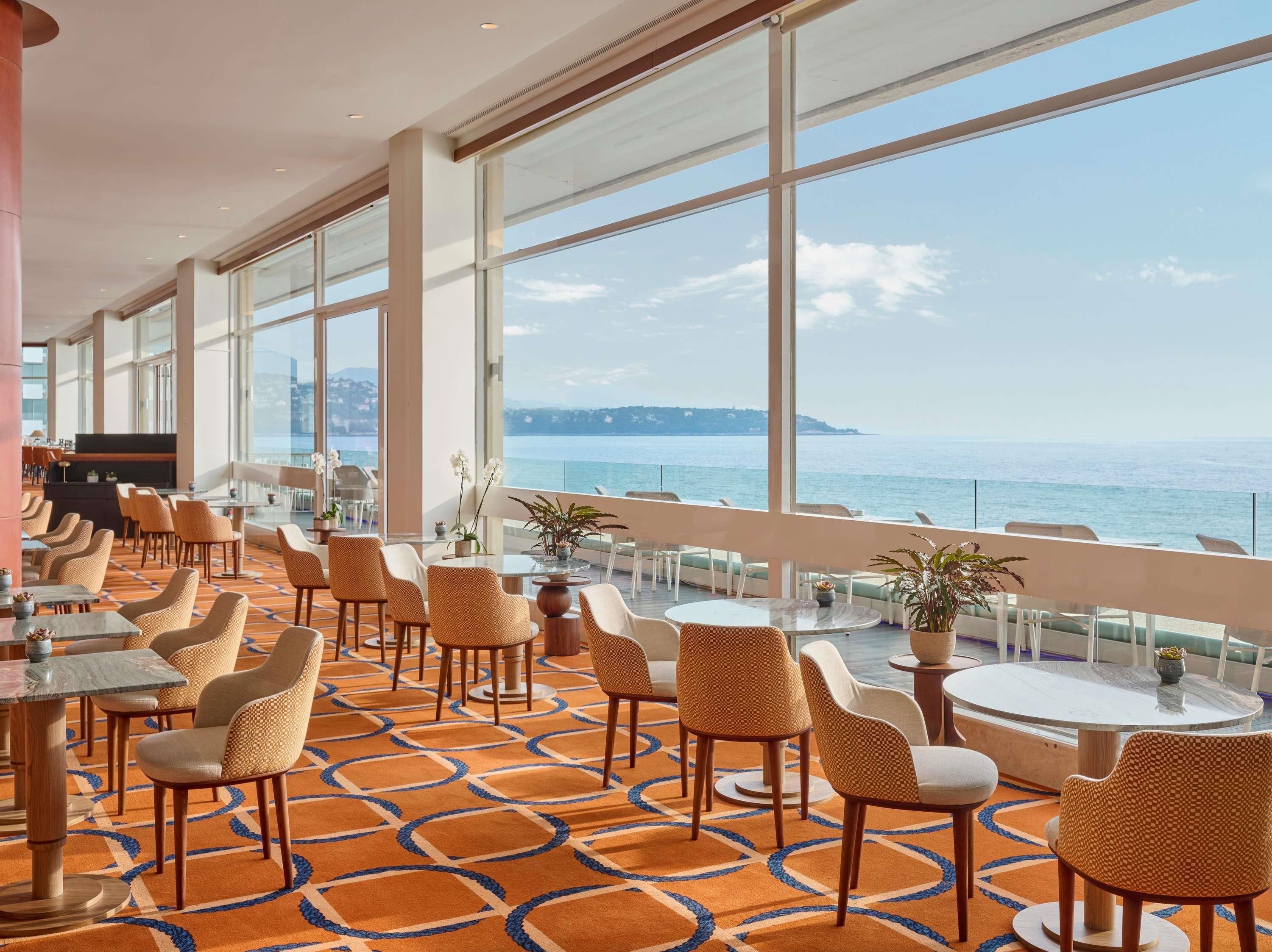 Un hall de hotel con un amplio ventanal con vistas al mar, amueblado con sillas y mesas de mimbre, y una zona de bar.