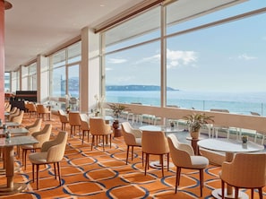 Un hall de hotel con un amplio ventanal con vistas al mar, amueblado con sillas y mesas de mimbre, y una zona de bar.