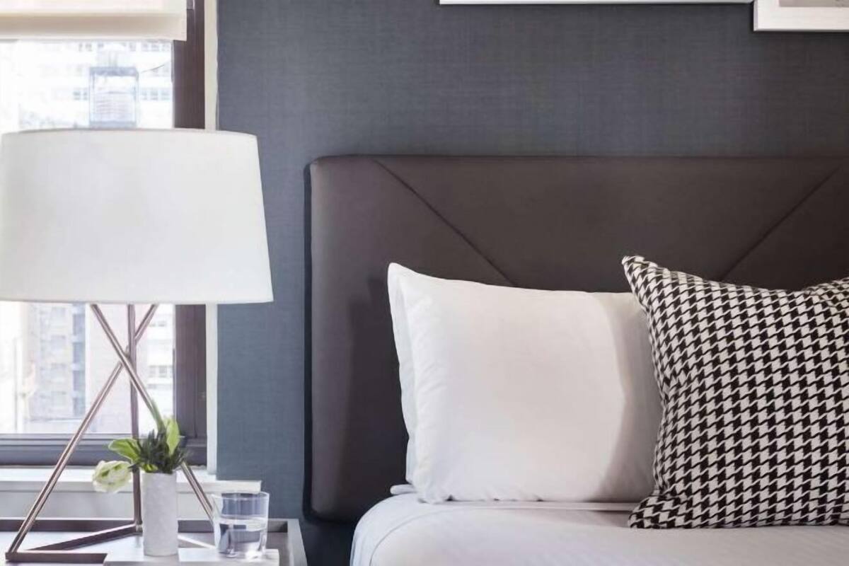 Une chambre d’hôtel avec un lit, une lampe de chevet, une plante en pot et une fenêtre donnant sur des immeubles.