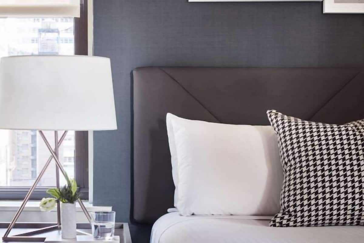 Habitación de hotel con cama, lámpara de noche, planta en maceta y ventana con vista a edificios.