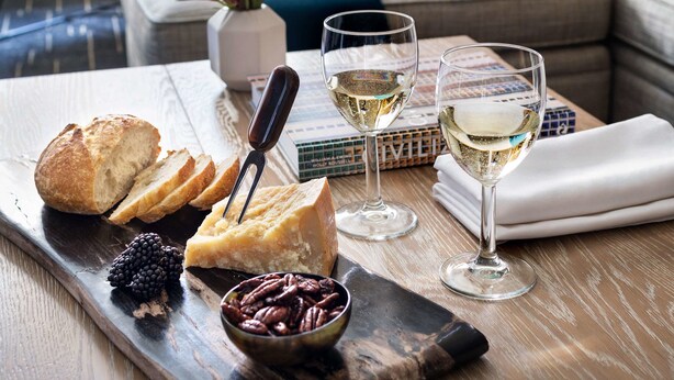A wooden board with sliced bread, cheese, blackberries, and pecans, accompanied by two glasses of white wine, set on a table in a room with a view of buildings through a window.