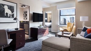 A modern hotel room with a desk, a sofa, a chair, a flat-screen TV, and a large window with a city view.