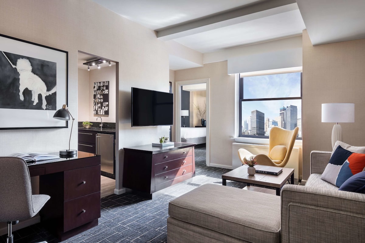 Quarto de hotel moderno com escrivaninha, sofá, cadeira, TV de tela plana e uma grande janela com vista para a cidade.