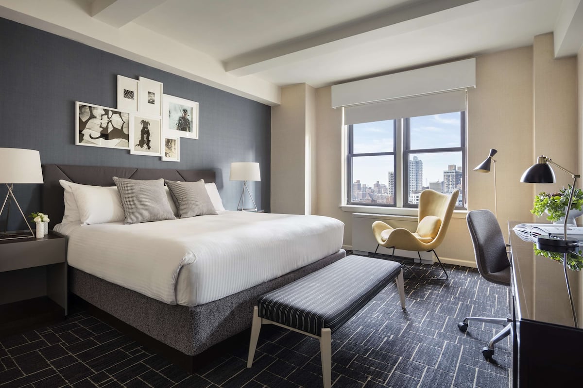 A modern hotel room with a large bed, a desk, a chair, and a window with a city view.