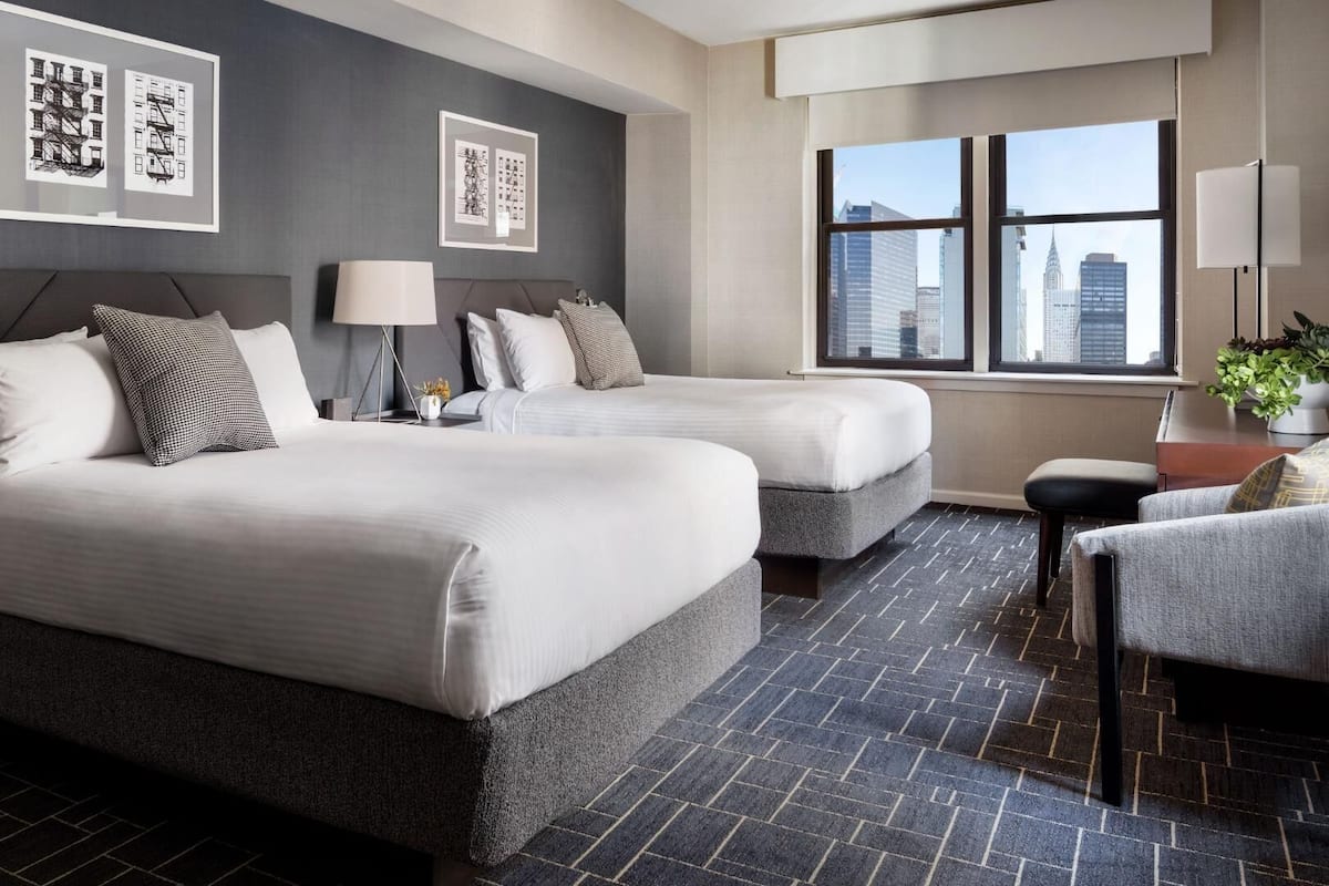 A hotel room with two beds, a desk, a chair, a window with a city view, and artwork on the walls.