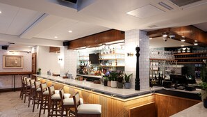 A bar area with wooden bar stools, a counter with a cash register, and shelves stocked with bottles and glasses.